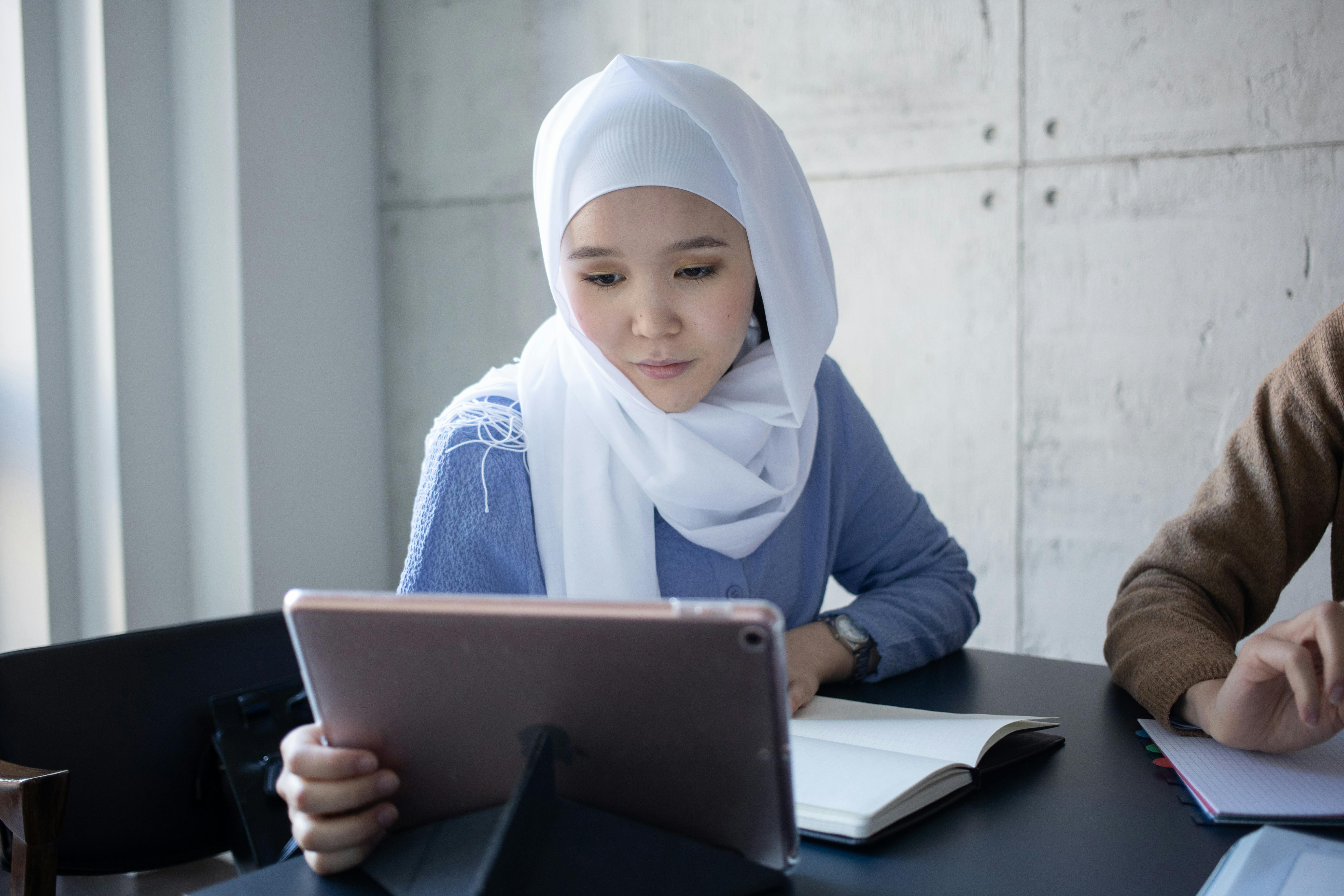 Student at a desk focusing on a tablet with a notebook open beside her in a bright study space. Photo: Gabby K / Pexels.
