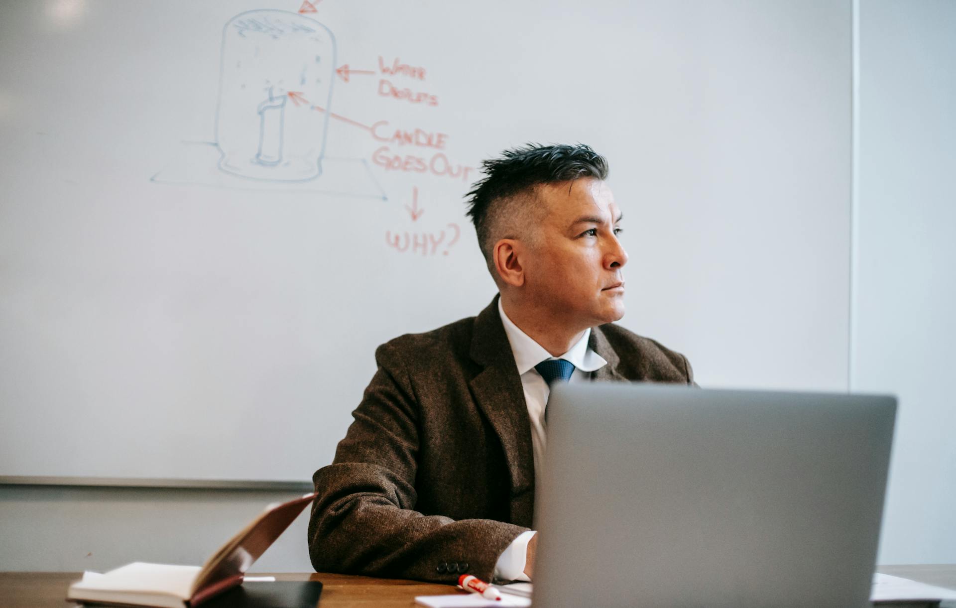Educator at a desk with a laptop, whiteboard with diagrams behind. Photo: Vanessa Garcia / Pexels.