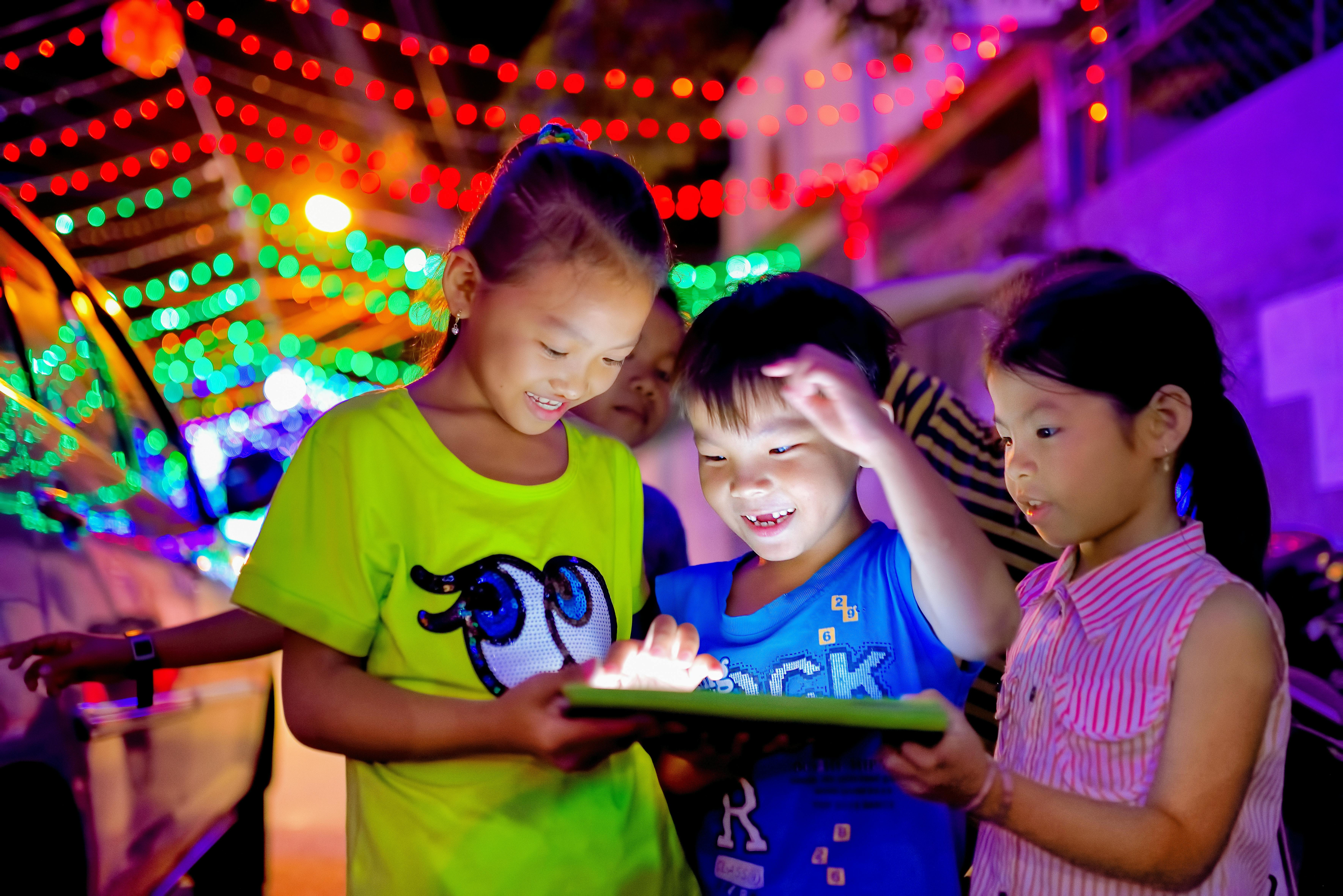 Children gathered around a glowing tablet outdoors at night with colourful lights. Photo: Vi N Dong / Pexels.
