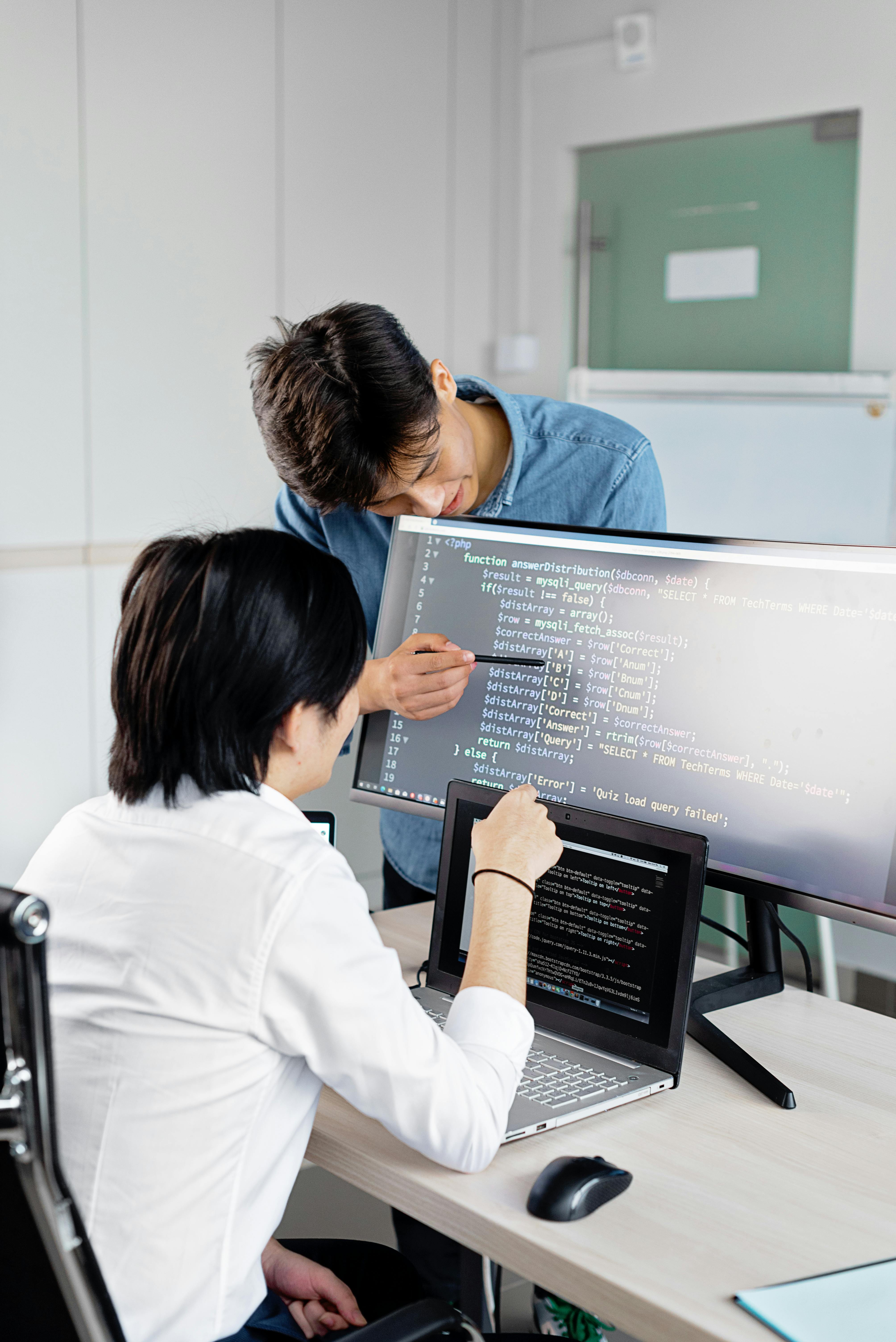 Two students pair-programming at a desktop monitor and laptop in a computer lab. Photo: Mikhail Nilov / Pexels.