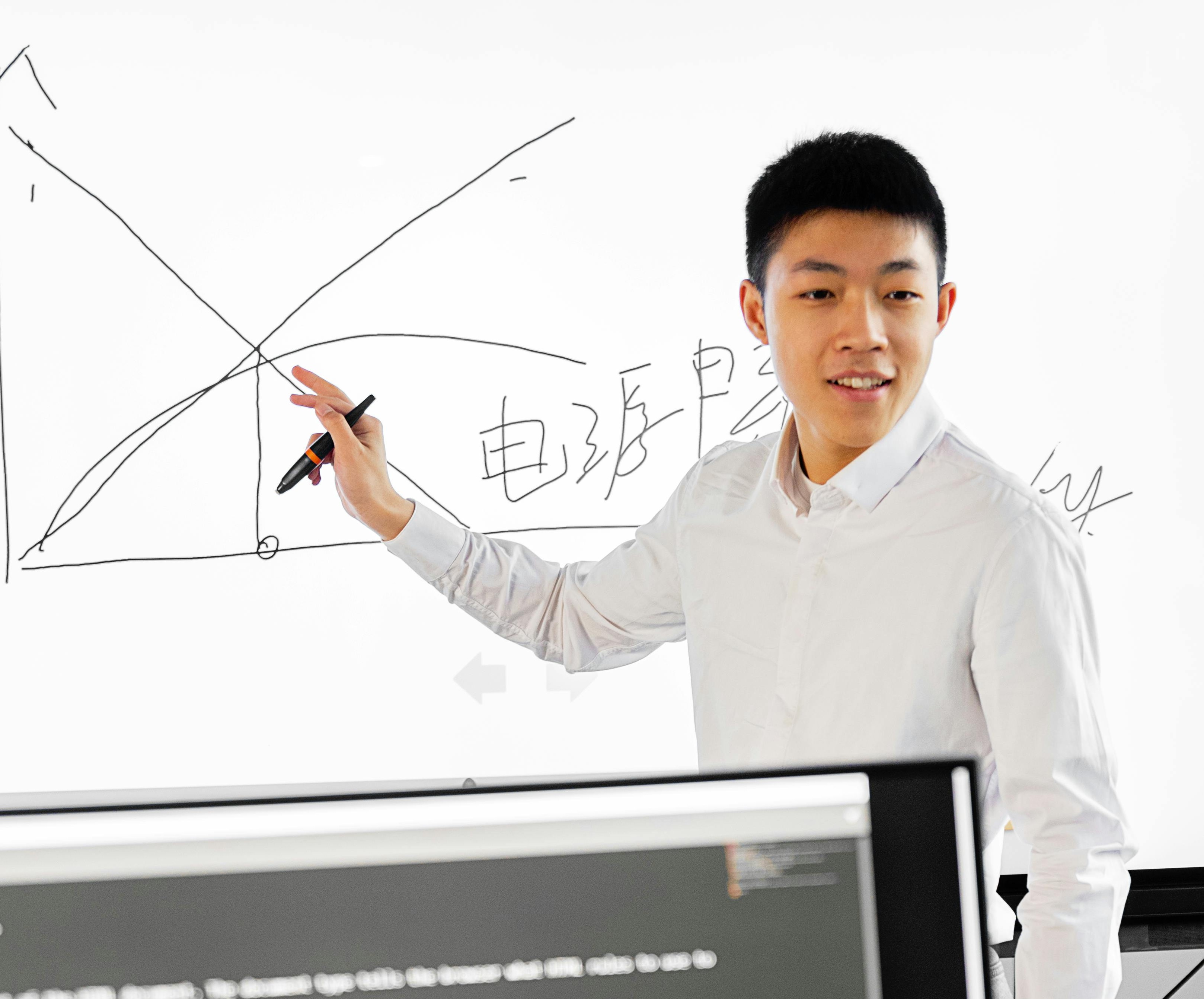 Student at a whiteboard with physics diagrams and Chinese labels, with coding visible on monitors in the foreground. Photo: Mikhail Nilov / Pexels.