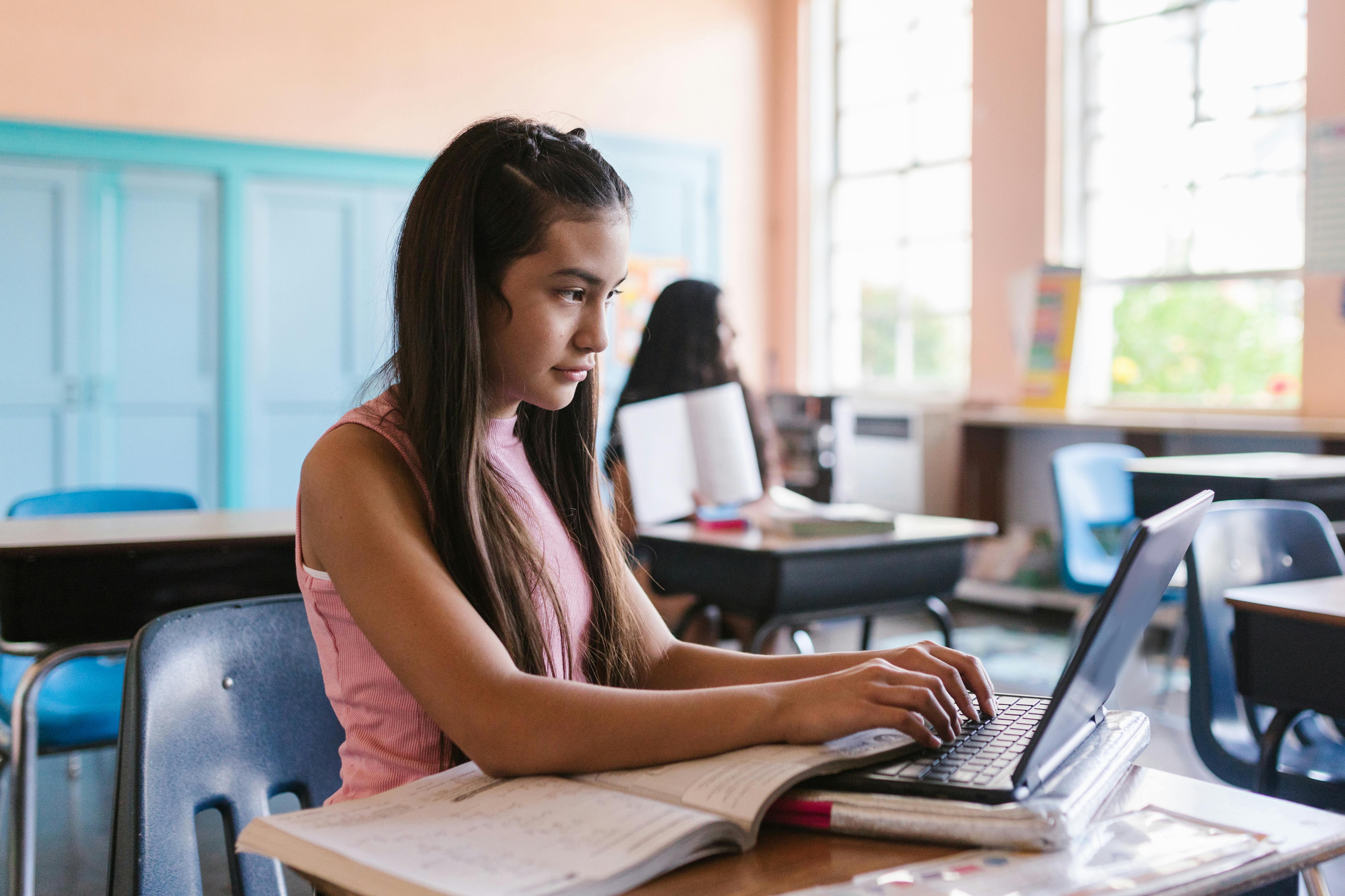 Student using a laptop alongside a textbook in a bright classroom. Photo: RDNE Stock project / Pexels.